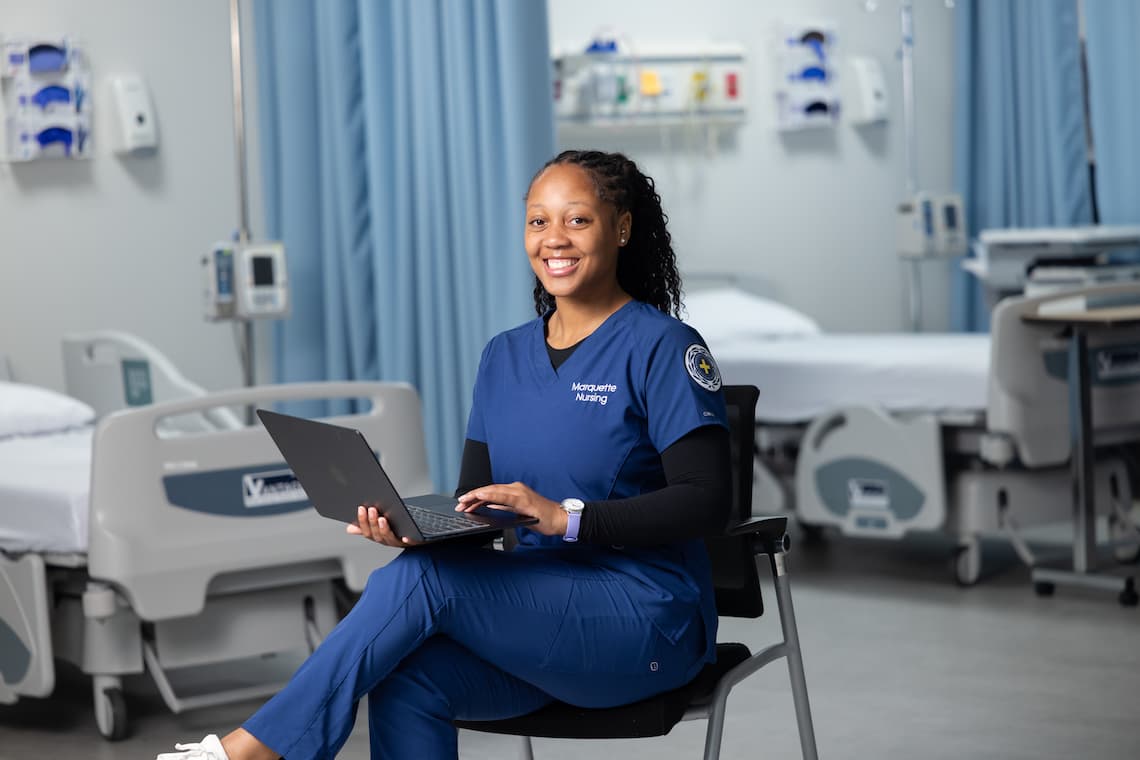 Nursing student sitting in a chair holding their laptop and smiling
